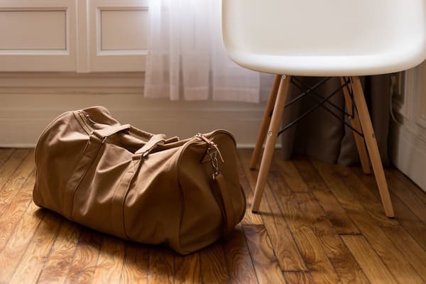 A tan duffle bag on wood floors next to a white chair