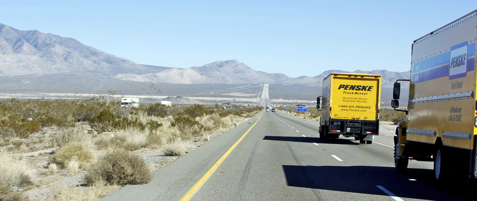 A Penske truck towing a car on a long flat road
