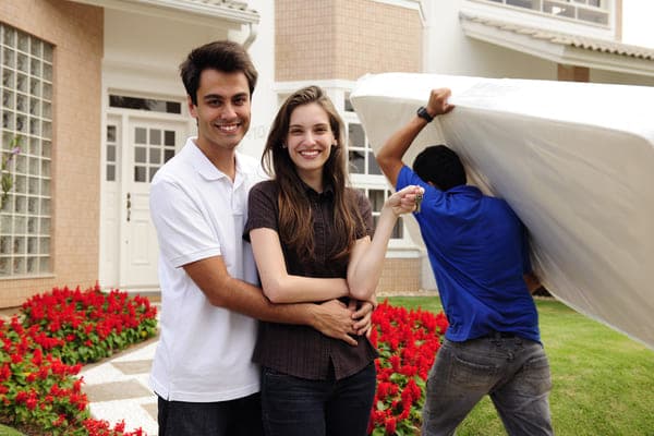 Two new homeowners stand in front of their home as a mover carries a mattress inside