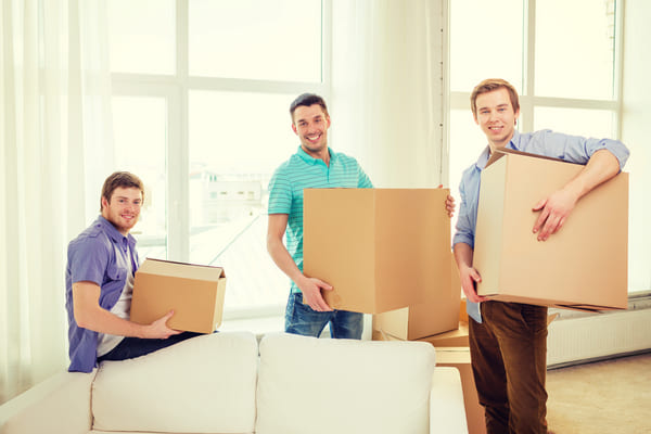 three young men stand in a living room holding moving boxes